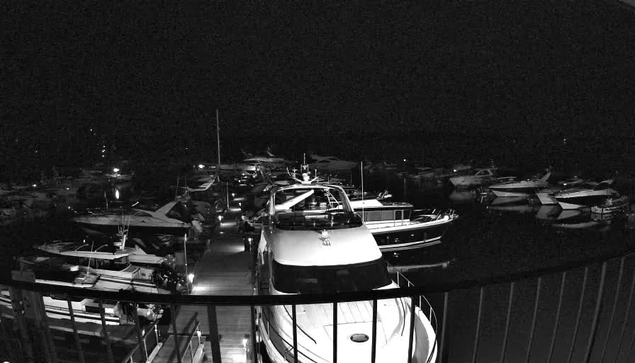 A dimly lit marina at night, featuring numerous boats docked closely together. The foreground shows a large white boat with a prominent bow and a railing, alongside smaller vessels. The dark water reflects faint lights from the boats and surrounding areas, creating a serene but shadowy atmosphere. The overall scene lacks bright colors due to nighttime conditions.