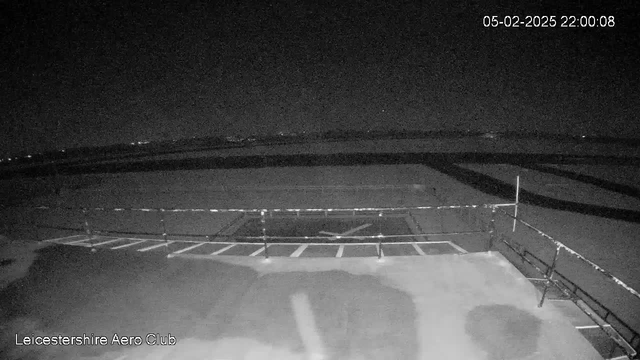 A dark scene captured from a webcam shows an empty airport tarmac at night. The ground is mostly dark with some indistinct shapes, and faint outlines of a runway in the distance. A thin white line runs along the edge, possibly indicating a parking area. The date and time, 05-02-2025 22:00:08, are displayed in the top right corner. There are no visible vehicles or people in the scene.