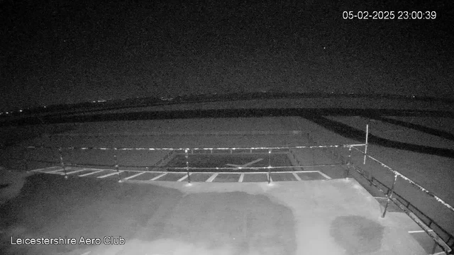 A nighttime view from a webcam at the Leicestershire Aero Club shows a dark, empty tarmac area with a fence visible in the foreground. The sky is mostly dark with minimal light, possibly from distant buildings or stars. There are faint outlines of runway markings on the ground, and the date and time are displayed in the top corner.