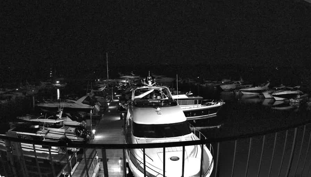 A nighttime scene of a marina filled with various boats docked side by side. The image is mostly dark, with a few illuminated areas highlighting the white hull of a large yacht in the foreground and smaller boats further back. Reflections of the boats are faintly visible in the water, and there are faint lights from nearby structures and boats creating a subtle glow in the otherwise dark scene. The perspective suggests a viewpoint from a balcony or dock overlooking the marina.