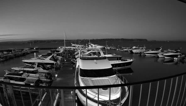 A black and white image of a marina filled with various boats and yachts docked in the water. The scene is taken from a viewpoint overlooking the marina, with a wooden pier visible in the foreground. The background features calm water and a distant landmass. The overall atmosphere is tranquil, with clear skies and the silhouettes of boats creating a serene setting.