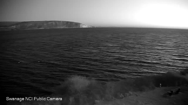 A black and white image of a coastline with a beach in the foreground and waves gently lapping at the shore. The ocean extends to the horizon, where cliffs are visible in the distance under a lightening sky, suggesting dawn or dusk. There are two benches on the sandy area, and the scene is calm and serene, with gentle ripples across the water's surface.