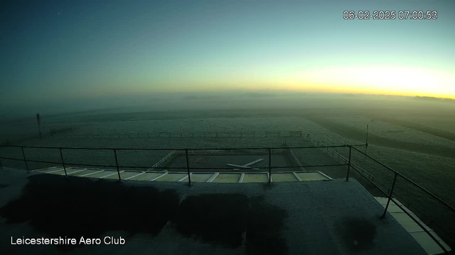 A foggy landscape is visible in the image, taken at dawn. The horizon shows a gradient of soft yellow and blue colors as the sun rises. In the foreground, there is a flat rooftop with a railing and a small airplane symbol marked on the surface. There are blurred outlines of trees and fields in the background, partially obscured by fog. A windsock stands to the left, indicating wind direction. The date and time are displayed in the top right corner.