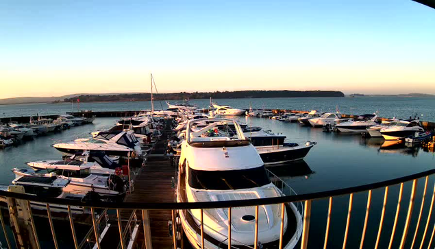 A scenic view of a marina at sunset. There are numerous boats and yachts docked in the water, reflecting the warm hues of the setting sun. A wooden dock runs alongside the boats, and a railing is visible in the foreground. The sky above transitions from soft blue to warm orange, with distant hills visible across the water.