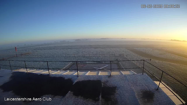 A view from a webcam at Leicestershire Aero Club shows a frost-covered airstrip in the early morning. The horizon is hazy with mist, and the sky is clear, transitioning from dark blue to light blue as the sun rises. A windsock, visible on the left, indicates the wind direction. The airstrip is bordered by a railing, and there is a light-colored airplane silhouette on the tarmac. The overall scene conveys a calm, tranquil atmosphere.