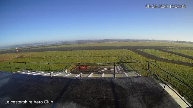 A clear blue sky is visible above a green field. In the foreground, a dark surface with railings runs along the edge, and a section of pavement leads to a helipad marked with white lines. The field is bordered by wooden fences, and in the distance, a variety of runways can be seen. There are no clouds in the sky, and the scene is bright and sunny. The image is timestamped with the date and time at the top right corner.