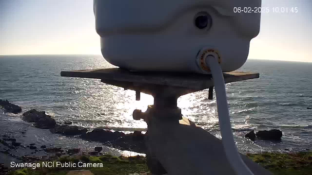 A white cylindrical object is at the top of the image, mounted on a weathered wooden platform. Below it is a sloped metal bracket. In the background, the sunlight reflects off a calm sea, which stretches to the horizon. The shoreline features dark rocks and some greenery in the foreground. The sky is clear with a slight glow from the sun. A timestamp in the top right corner indicates the date and time.