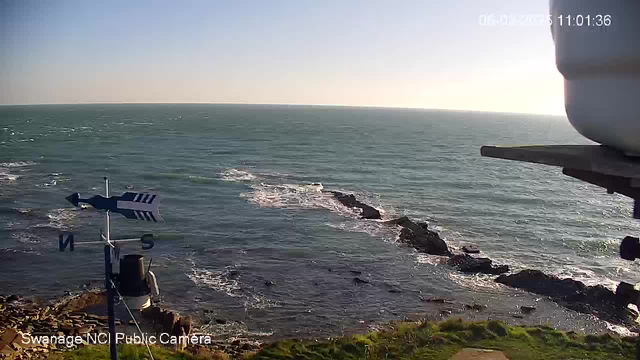 A coastal view shows the turquoise sea with gentle waves lapping at rocky outcrops. There's a grassy area in the foreground, and a weather vane with directional markings on the left side of the image. The sky is clear and blue, indicating a bright day, while the sunlight reflects off the water, creating a shimmering effect. The timestamp at the top right shows the date and time.