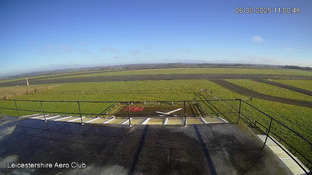 A clear blue sky is visible above a grassy landscape with a large open field. In the foreground, a flat rooftop with railings features markings resembling a helicopter landing pad. Beyond, there is a long runway leading into the distance, bordered by a wooden fence. The scene is bright and sunny, indicating daytime. The date and time displayed in the corner reads 06-02-2025 at 11:00:48.