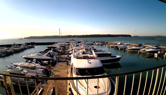 A marina scene with several boats docked in calm water. The boats vary in size and design, with some larger vessels prominently displayed. A wooden dock lines the marina, leading to the boats. In the background, a calm sea meets a clear sky, with hints of land in the distance. The sunlight casts a warm glow over the water and boats, creating a serene atmosphere.