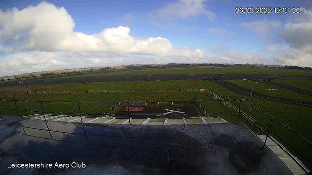 A wide view from a webcam at Leicestershire Aero Club, showing a grassy airfield under a partially cloudy sky. The ground is divided into sections of green grass and dark pavement. In the foreground, there is a white railing and a section of the building's roof. The runway is visible with a red flag on one side, and a clear view stretches toward distant hills under a blue sky with scattered clouds. The image captures a peaceful, open landscape.