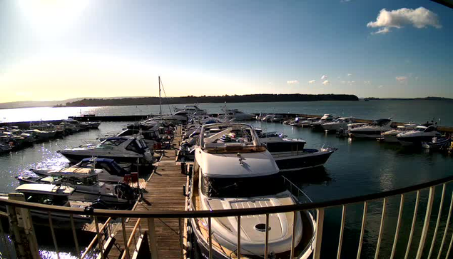 A view of a marina on a sunny day with several boats docked in the water. The scene includes a wooden dock lined with various boats, some larger yachts and smaller vessels, reflecting light from the water. The background features a calm body of water leading to distant land under a clear blue sky with a few scattered clouds. The overall atmosphere is peaceful and inviting.