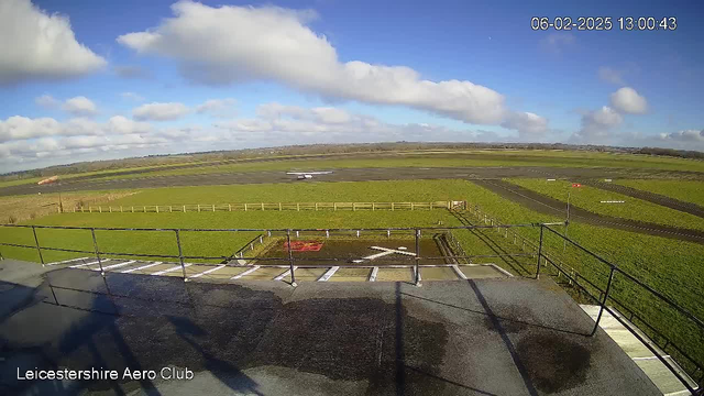 A landscape view from the Leicestershire Aero Club webcam captures a wide, open grass field under a partly cloudy blue sky. In the foreground, a tarmac area with a white marked "X" is visible, alongside a railing. A small aircraft is seen on the runway to the left, while a wooden fence lines the edge of the grass field. The rest of the land stretches out in the background, blending into a distant horizon.