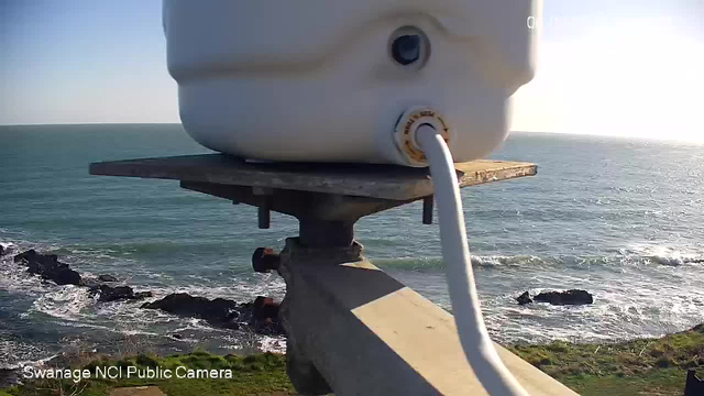 A close-up view of a white cylindrical container mounted on a metal platform. The container has a pipe running from it. In the background, the ocean stretches out to the horizon, with waves gently lapping against rocky shoreline. The sky is clear and blue, with a bright light indicating sunlight in the scene.