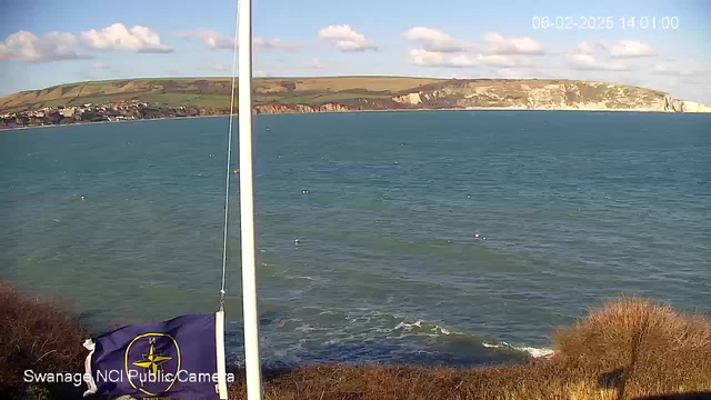 A coastal scene captured by a webcam, showing calm blue water reflecting the sky. In the foreground, there is a flag with a nautical design flapping gently in the breeze. To the left, there is a shoreline with buildings dotting the landscape. In the background, grassy hills rise, leading to a rocky cliff that meets the ocean. The sky is mostly clear with a few scattered clouds, and the timestamp indicates the image was taken in February 2025 at 2:01 PM.