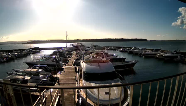 A harbor scene is depicted with numerous boats and yachts docked in the water. The sun is shining brightly, reflecting off the surface of the water, creating a sparkling effect. In the background, there are hills and a clear blue sky, with a few clouds scattered. A wooden dock extends into the harbor, bordered by a railing. The boats are of various sizes, some are larger yachts, while others are smaller vessels.