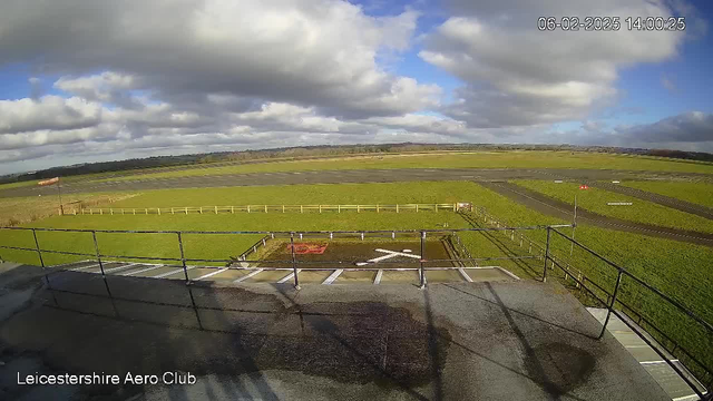 A wide view of an airfield at the Leicestershire Aero Club, featuring a green grassy area with a black runway in the foreground. In the distance, there are white wooden fences bordering the airfield. The sky is partly cloudy, with bright blue patches visible. Time and date are displayed in the corner, indicating February 6, 2025, at 14:00. Sockets and markings on the ground are visible near the edge of the airstrip.