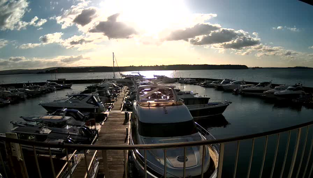 A marina filled with numerous boats docked at the waterfront, with a wooden walkway leading to them. The sun is setting in the background, casting a golden light across the water. Clouds are scattered in the sky, creating a picturesque scene of a tranquil evening by the harbor.