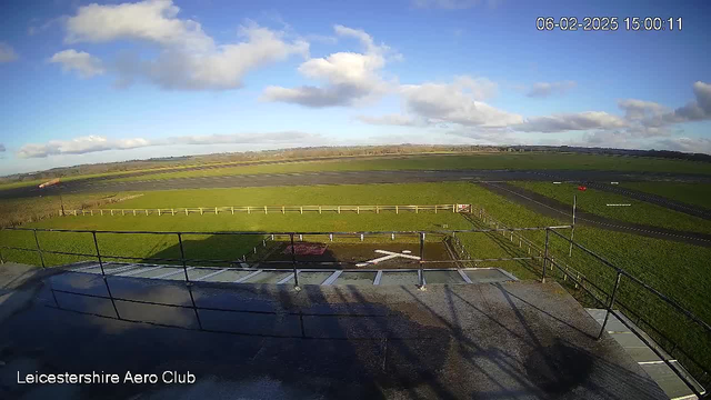 A clear view of a grassy airfield under a blue sky with scattered clouds. In the foreground, a railing is visible, suggesting a viewing platform. There is a white cross marking on the ground, indicating a possible helipad. Beyond the cross, the field extends with a few trees in the distance, and a runway is visible on the right where a red windsock is positioned. The image captures a bright, sunny day.