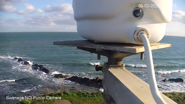 A white circular object, possibly a camera, is mounted on a gray platform that is attached to a metal pole. Below, the ocean waves crash against a rocky shoreline. The water is a deep blue, with white foam at the wave crests. In the background, there are scattered clouds in a light blue sky. The foreground has a patch of green grass near the base of the pole. A small section of black rocks is visible in the water.