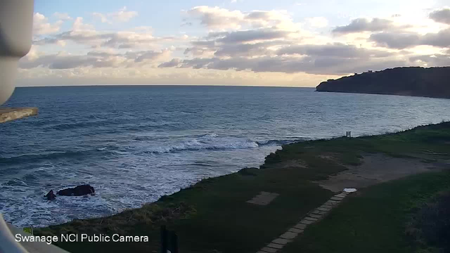 A coastal scene showing rolling waves in the ocean under a partially cloudy sky during sunrise or sunset. The foreground features grassy land leading to the water, with a path made of stone slabs leading toward the shore. A large rock is visible in the water, and a steep cliff can be seen in the background to the right. The overall atmosphere conveys a calm and serene environment.