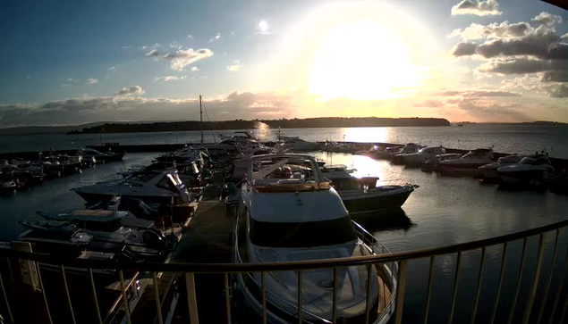 A marina at sunset with numerous boats docked. The sun is low in the sky, casting a warm glow over the water. There are fluffy clouds scattered in the sky, and the shore outlines the background. The scene evokes a calm and serene atmosphere.