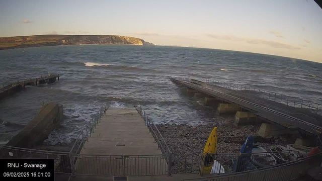 A scenic view of a coastal area with a rocky shore and choppy waves crashing against it. In the foreground, there is a concrete pathway leading down to the water, bordered by a metal railing. To the right, some small boats are partially visible, including a yellow kayak. In the background, steep cliffs rise above the sea, with a clear sky and a few clouds overhead, indicating late afternoon. The overall ambiance suggests a breezy, lively marine environment.