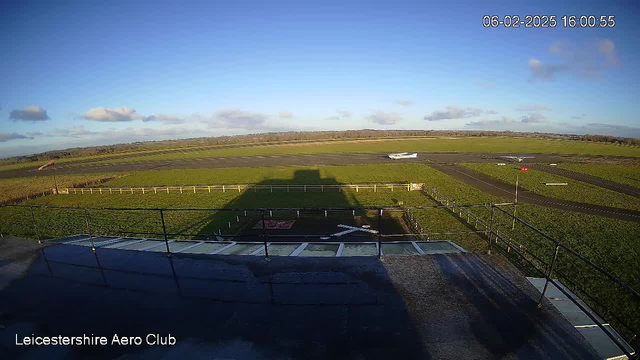 A view from a webcam at Leicestershire Aero Club. The image captures a clear blue sky with scattered clouds. In the foreground, a runway stretches out, bordered by green grass. A small white airplane is positioned on the runway to the right. The shadow of the observation tower is cast on the ground. A nearby fence separates the runway from the grass, and the time stamp indicates the image was taken at 16:00 on February 6, 2025.