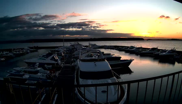 A view of a marina at sunset, with several boats moored along the water. The sky is filled with clouds in shades of orange, pink, and gray, reflecting on the calm water surface. The silhouette of a distant shoreline is visible in the background. A railing in the foreground adds depth to the scene.