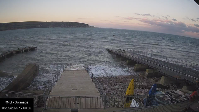 A coastal view showing a stone pathway leading down to the water, with a metal railing on either side. Small waves are visible on the water's surface. To the right, there are two wooden piers extending into the sea at different angles. A rocky shoreline is on the left, with colorful kayaks (yellow, blue, and red) stacked nearby. The sky has soft clouds, and a faint sunset glow is visible in the distance.