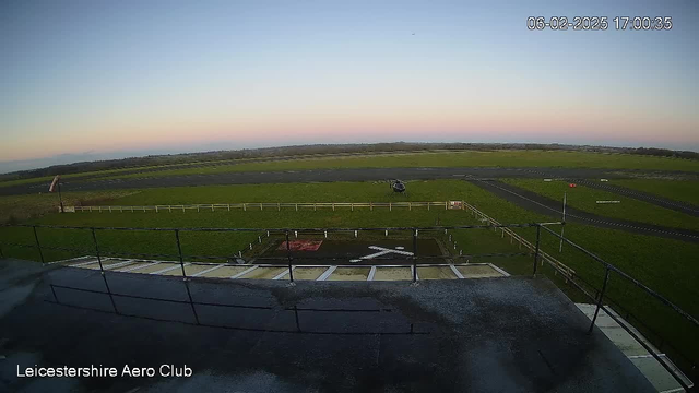 A view from a webcam positioned on top of a building at Leicestershire Aero Club. In the foreground, there is a rooftop railing and part of the building’s surface. Below, an open green field stretches out with a large, flat runway. A helicopter is visible on the runway, surrounded by white fencing. The sky shows a gradient from blue to light pink, indicating early evening. The image captures the serene landscape and aviation setting, with the date and time displayed in the top right corner.