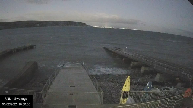 A view of a shoreline at dusk, with a dark sky and clouds. The water appears choppy, reflecting some light. In the foreground, there is a rocky area leading to a wooden pier on the left, and a second shorter pier extending into the water. To the right, there are several kayaks in various colors, including yellow and blue, stored near a railing. The landscape in the background features a distant shoreline or cliffs. The setting suggests a coastal area, possibly a harbor or beach.