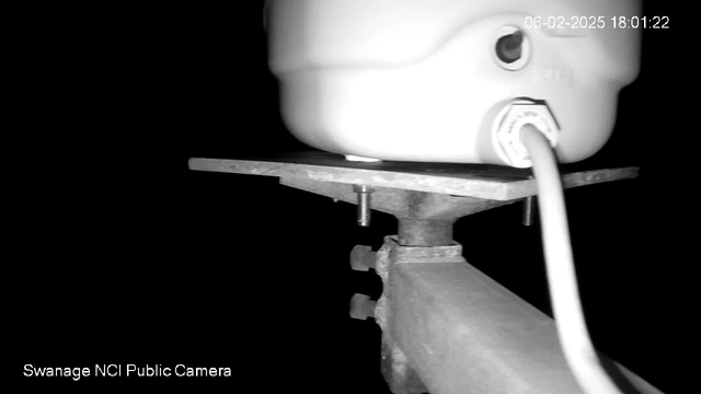 A close-up view of a white, cylindrical object resting on a metal plate. The plate is supported by a metallic structure with visible bolts and a power cable connected to the object. The background is dark, indicating low light conditions. A timestamp is visible in the top right corner, indicating the date and time.