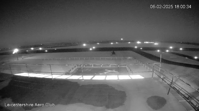 A dark, nighttime image from a webcam showing an airport or aerodrome environment. In the foreground, there is a rectangular helipad marked with an "X" on a dark surface. The background includes a runway and several distant lights, possibly from vehicles or buildings. The overall scene appears dimly lit with a cloudy sky, indicating it’s nighttime. The date and time displayed at the top right corner reads 06-02-2025 at 18:00:34. A label in the bottom left indicates the location as "Leicestershire Aero Club."