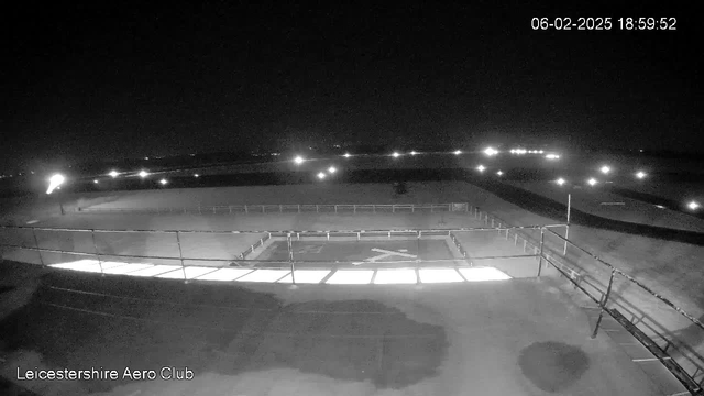 A nighttime view from a webcam at the Leicestershire Aero Club, showing a low-lit area with a fenced platform. The central part displays a light-colored landing area with an 'X' shape marked on it. Surrounding the landing area are faint lights from the distance, illuminating parts of the scene. The image is predominantly dark, with limited visibility of surrounding features.