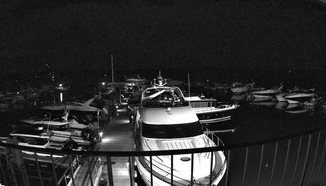 A nighttime view of a marina filled with various boats and yachts. Some boats are docked closely together, while others are moored further away. The scene is illuminated by soft lights reflecting off the water, creating a calm ambiance. A wooden walkway runs between the boats, with railings visible in the foreground, framing the scene.