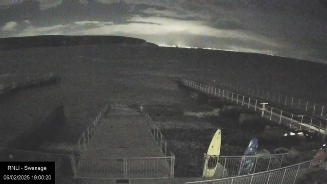 A dark landscape with a distant view of a coastline under a cloudy sky. The foreground shows a pathway leading to the water, bordered by metal railings. Two kayaks, one yellow and one blue, are secured near the pathway. The water appears still and the overall scene is dimly lit with hints of light reflecting in the distance.