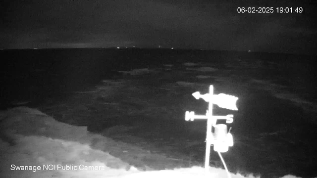 A nighttime webcam view showing tumultuous waters. In the foreground, a weather vane is visible, indicating wind direction with "N," "E," "S," and "W" markers. The dark ocean stretches out towards a horizon with faint distant lights. The image is dim, capturing a sense of a stormy sea under a cloudy sky. The date and time are displayed in the upper right corner.