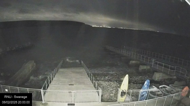 A dark scene depicting a shoreline at night. There is a narrow, concrete path leading down to the water, surrounded by railings. To the right, several kayaks are stacked on the ground, with one yellow kayak prominently displayed. The water is visible but mostly obscured by shadows, and the distant shoreline is faintly illuminated by city lights. The sky is overcast and dark. The date and time are displayed in the bottom left corner.