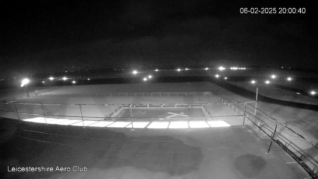 A dark, nighttime scene captured by a webcam at Leicestershire Aero Club. The image features a flat, open area with faint lights in the distance. A white cross shape is visible on the ground, illuminated by nearby lights. There are railings along the edge of the area, and a few scattered lights are seen beyond the perimeter, contributing to the low-light ambiance.