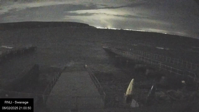 A dark scene depicting a pier extending into the water, with faint silhouettes of boats on the shoreline. The sky is overcast, and there are low clouds obscuring the moonlight, creating a dimly lit atmosphere. In the background, hills can be seen against the horizon. The pier is partially illuminated, with some details of the wooden railing visible.