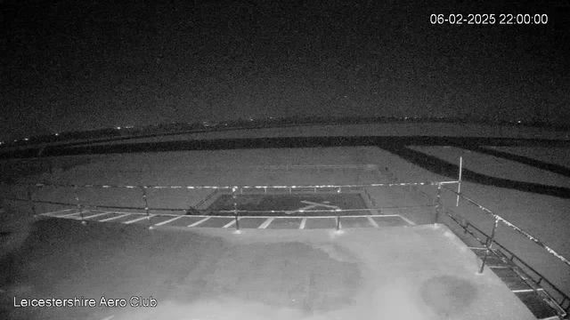 A dark webcam image showing a runway with a fence in the foreground. The ground is mostly flat with markings indicating parking spaces. In the background, there are faint lights suggesting distant structures or roads under a nighttime sky. The timestamp in the top right corner indicates it is 10:00 PM on February 6, 2025. The bottom left corner has the text "Leicestershire Aero Club."