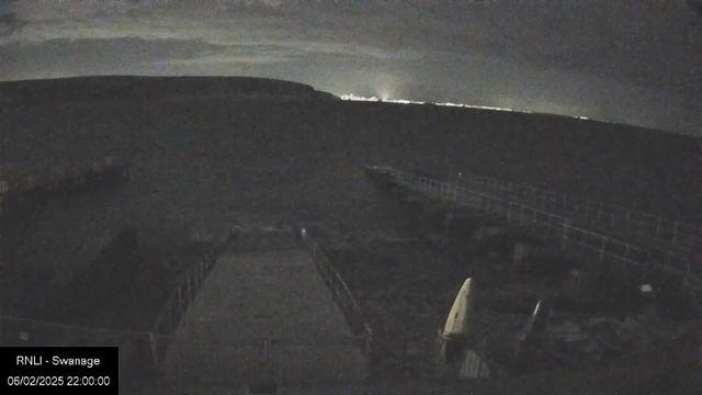 A dimly lit scene captured at night showing a wooden walkway leading to the water. In the foreground, there are several objects that appear to be boats or kayaks, with one white kayak prominently displayed. The background features a dark hillside against a cloudy sky, with faint lights from a distant area hinting at a town or city. The image has a grainy texture, emphasizing the low light conditions.