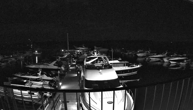 A nighttime scene of a marina with several boats docked. The image is predominantly in black and white, highlighting the silhouettes of the boats. In the foreground, a large boat is positioned directly in front, while smaller boats are lined up to the sides. Reflections of lights can be seen in the water, with some distant lights illuminating parts of the marina. The overall atmosphere is calm and serene, with shadows and limited visibility.