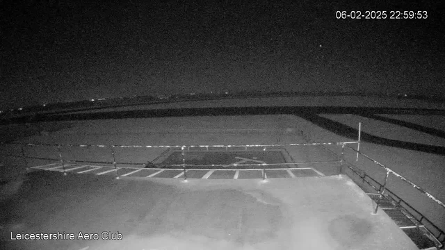 A dark scene captured from a webcam at night, showing a runway and surrounding area. A faint outline of the runway is visible, with some horizontal lines indicating markings. A railing runs across the bottom of the image. The time and date in the top right corner show it is February 6, 2025, at 22:59:53. There are distant lights in the background, possibly from nearby buildings or vehicles, and the sky is mostly dark with a few stars visible.