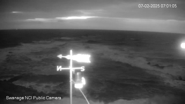 A dark, monochrome webcam image showing a turbulent sea under a cloudy sky. The horizon is slightly visible, with waves crashing in the foreground. A wind direction indicator with a compass rose is positioned in the lower left, pointing north, with the letters 'N' and 'S' clearly marked. The timestamp in the top right indicates the date and time as July 2, 2025, at 07:01:05.