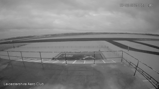 A black and white image showing a wide view from an elevated position at Leicestershire Aero Club. The ground is mostly empty with a large, clear runway extending into the distance. There is a faint outline of distant hills in the background under a cloudy sky. In the foreground, a fenced area with a large "X" marked inside suggests a landing or takeoff area, and a viewing platform rails can be seen along the lower part of the image. The timestamp in the corner indicates the date and time as 07:00:46 on February 7, 2025.