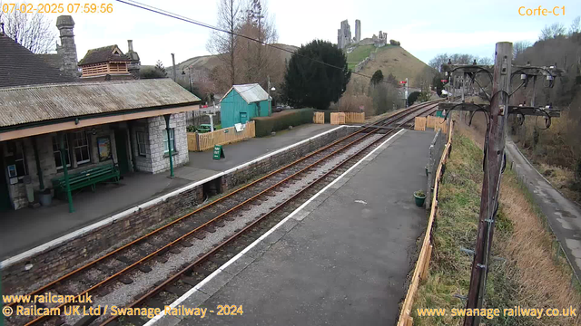 A view of a railway station platform with stone buildings and a wooden-roofed structure on the left. A green bench is situated on the platform, and a sign reading "WAY OUT" is visible on the ground. Two railway tracks run parallel along the bottom of the image, with gravel between them. In the background, rolling hills rise, with an ancient castle visible on a hilltop. Trees and shrubs are present on the hillsides, and there is a wooden fence to the right.