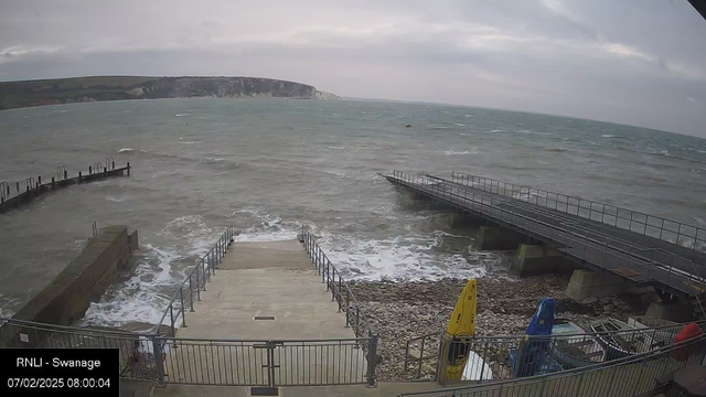 A view of a coastline with choppy waves under a cloudy sky. In the foreground, there are stairs leading down to the water, surrounded by metal railings. To the right, colorful kayaks are secured to the railing, and a wooden pier extends into the water from the left side. The cliffs can be seen in the distance. The scene appears overcast and windy.