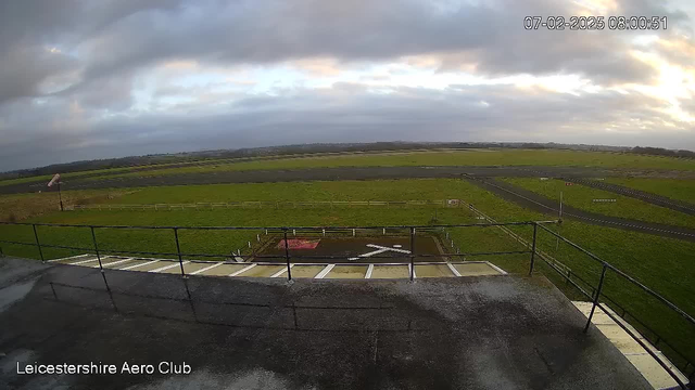 A view from a high vantage point overlooking a grassy airfield. The sky is mostly cloudy with some breaks of light. In the foreground, there is a railing along the edge of a flat rooftop. Below, a small section of the airfield features a marked area with an 'X' symbol and a red square. There are dirt paths and visible boundaries separating sections of grass. A wind direction indicator is present, and a road runs alongside the airstrip, leading toward the horizon.